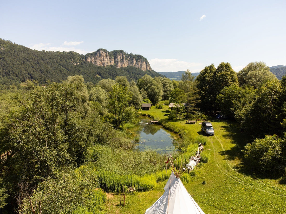 Idyllischer Campingplatz mit Van, Teich und Bergen, umgeben von grüner Natur im Sommer.